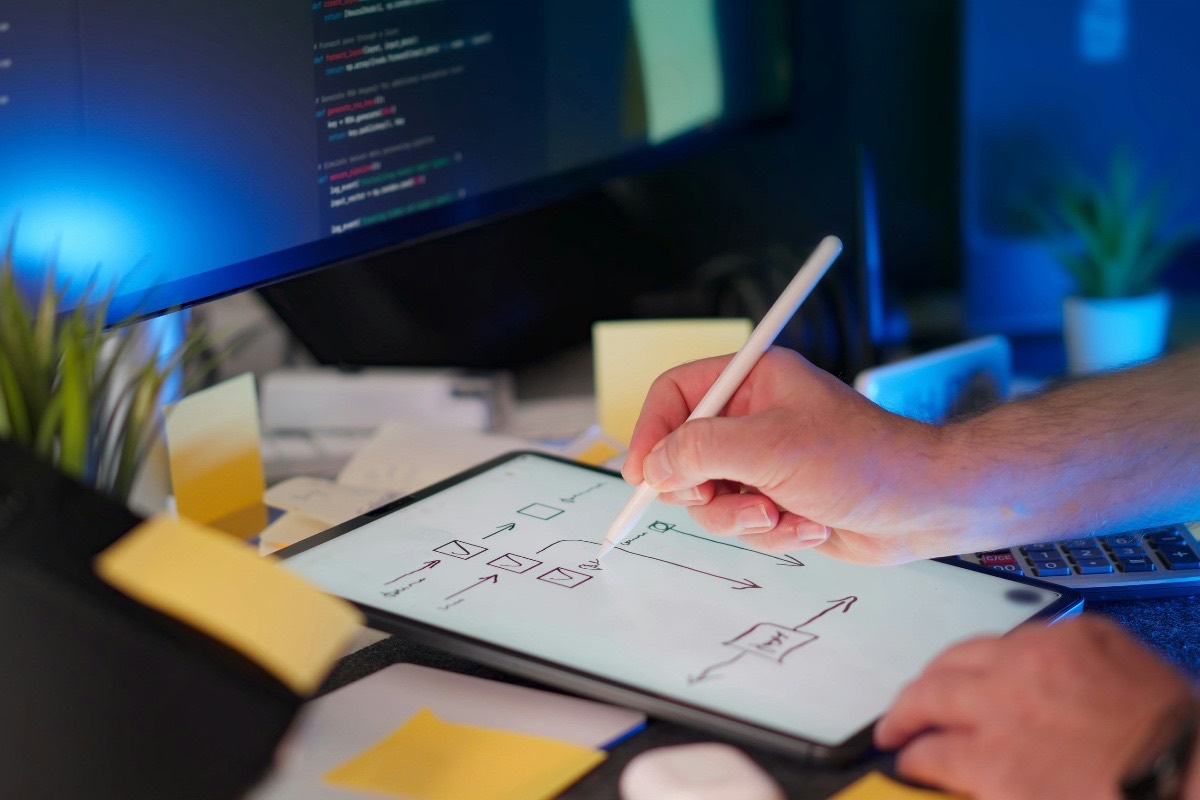 Developer sketching a system architecture diagram on a tablet beside a monitor showing code